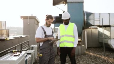 Tulumlu, gülen sakallı bir işçi, elinde modern tabletle siyahi bir mühendisle el sıkışıyor. Çok kültürlü insanlar, tesisin çatısında başarılı bir işbirliği içinde olmaktan memnuniyet duyuyorlar..