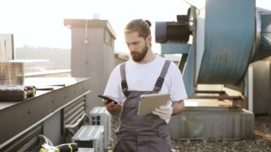 Tulum giymiş sakallı usta ayakta dikilip tablet tutarken aynı zamanda dijital akıllı telefonu temiz havada kullanıyor. Alet kullanan ve fabrikanın çatısında teçhizat taşıyan beyaz yetenekli bir adam..