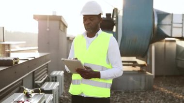 Ciddi bir fabrika işçisi fabrikadaki cihazları tamir ederken modern tableti elinde tutuyor. Afrika kökenli Amerikalı adam yerel çatıda çalışırken üniforma giyip teftiş yapıyor..