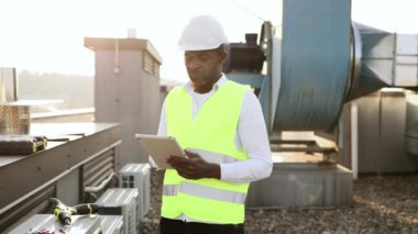 Beyaz şapka takan becerikli bir zanaatkar dışarıda elinde kablosuz tablet tutuyor. Cihazda bilgi arayan ve çatıdaki tesiste yapılan çalışmaları izleyen siyah konsantre adam.