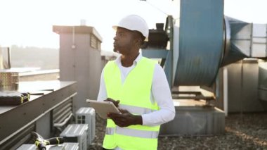 Ciddi bir fabrika işçisi fabrikadaki cihazları tamir ederken modern tableti elinde tutuyor. Afrika kökenli Amerikalı adam yerel çatıda çalışırken üniforma giyip teftiş yapıyor..