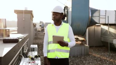 Ciddi bir fabrika işçisi fabrikadaki cihazları tamir ederken modern tableti elinde tutuyor. Afrika kökenli Amerikalı adam yerel çatıda çalışırken üniforma giyip teftiş yapıyor..