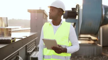 Beyaz şapka takan becerikli bir zanaatkar dışarıda elinde kablosuz tablet tutuyor. Cihazda bilgi arayan ve çatıdaki tesiste yapılan çalışmaları izleyen siyah konsantre adam.