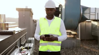 Beyaz kasklı ve yelekli inşaat işçisi ayakta dikilip temiz havada modern akıllı telefona bakıyor. Binanın çatısında fabrika ekipmanlarıyla çalışan zenci profesyonel bir adam..