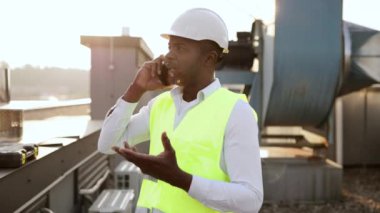 Temiz havada dijital cep telefonunda konuşurken ayakta durup gülümseyen iş üniforması giymiş erkek işçinin portresi. Afrikalı Amerikalı adam konuşuyor ve fabrikanın çatısında cihazlarla çalışıyor..
