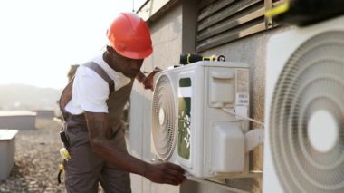 Yetişkin erkek üstat gri tulumlu ve turuncu kasklı klimaya eğiliyor ve havalandırmayı kontrol ediyor. Afrika kökenli Amerikalı bir adam binanın çatısını tamir ederken aletleri inceliyor..