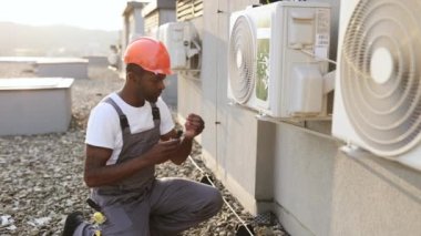 Afrika kökenli Amerikalı uzman bir tamirci, tek dizinin üstünde duruyor ve elinde tornavidayla binanın çatısında klima tamir ediyor. Fabrika bakımı kavramı.