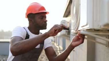 Odaklanmış Afrikalı bir zanaatkar, klimaya vida vidalarken elinde tornavidayla turuncu şapka takıyor. Ciddi bir uzman bozuk aygıtı tamir ediyor ve çatıda çalışıyor..