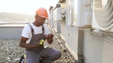 Gri tulumlu, turuncu şapkalı, modern tornavidayla birlikte klimanın yanında diz çökmüş yetenekli Afrikalı Amerikalı zanaatkârın yan görüntüsü. Onarım hizmeti kavramı.