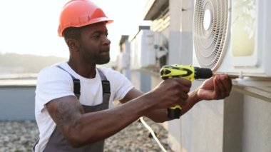 Dışarıdaki havalandırma sistemini tamir etmek için elektrikli tornavida kullanan yakışıklı Afro-Amerikan erkek portresi. Koruyucu kasklı gülümseyen adam doğrudan ve profesyonelce alet kullanıyor..