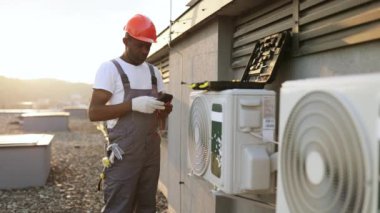 Gri tulumlu inşaat mühendisi elinde dijital çoklu metre tutuyor ve açık havada klima dağıtırken voltaj seviyesini kontrol ediyor. Afrika kökenli Amerikalı tamirci çatıya modern cihazlar yerleştiriyor..