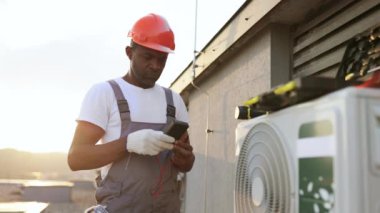 Konut binasının çatısına modern soğutma sistemi kurarken voltaj testini kullanan odaklanmış bir Afro-Amerikan uzmanı. Asılan klimada mevcut gücü ölçen yönetici.