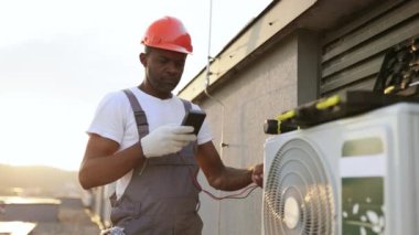 Konut binasının çatısına modern soğutma sistemi kurarken voltaj testini kullanan odaklanmış bir Afro-Amerikan uzmanı. Asılan klimada mevcut gücü ölçen yönetici.