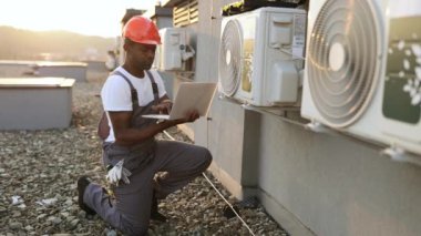 Klimanın yanında diz çökerken kablosuz bilgisayarda yazı yazan yetenekli bir adamın yan görüntüsü. Afrika kökenli Amerikalı uzman, soğutma sisteminin çabucak onarılması için modern teknolojileri kullanıyor..