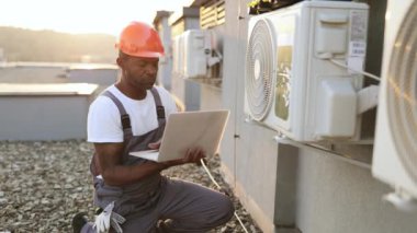 Sorumlu Afro-Amerikan işçi, taşınabilir dizüstü bilgisayarla çatıdaki soğutma tesislerini tamir ediyor. Arka planda güzel günbatımında klimanın yanında diz çöken meşgul bir adam..