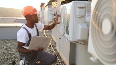 Sorumlu Afro-Amerikan işçi, taşınabilir dizüstü bilgisayarla çatıdaki soğutma tesislerini tamir ediyor. Arka planda güzel günbatımında klimanın yanında diz çöken meşgul bir adam..