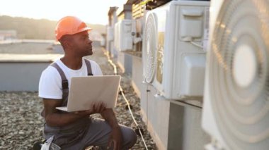 Çatıdaki klimanın yanında diz çökerken kablosuz bilgisayar kullanan konsantre erkek teknisyen. İş raporunda AC birimi hakkında modern cihazlar hakkında bilgi yazan Afrikalı Amerikalı uzman..