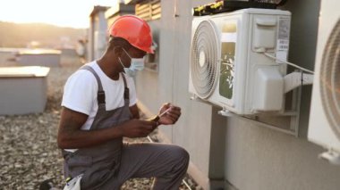 Koruyucu maskeli Afro-Amerikan tamircinin yan görüntüsü. Diz çöküp açık havada tornavidayla tamir ediyor. Yetenekli erkek, küçük arızalarla çabucak başa çıkıyor..