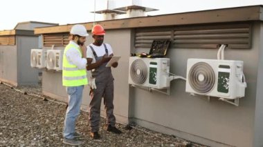 Elinde tabletle Afrika kökenli Amerikalı teknisyen çatıdaki beyaz şapkalı Kafkasyalı yöneticiye tamir edilmiş klimayı gösteriyor. Fabrika işçileri modern ekipmanları korumak için işbirliği yapıyorlar..