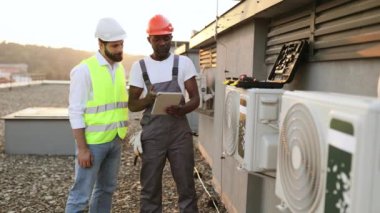 Çok kültürlü erkekler, açık havada bozuk soğutma sisteminin yanında dururken birbirlerine klima tamiri hakkında tavsiyelerde bulunuyorlar. Kullanıcı bilgili web sitelerini kaydırmak için tablet kullanan adamlar.