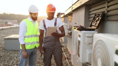Koruyucu şapkalı çok ırklı inşaat yöneticileri, klima tamiri hakkında modern tabletten bilgi okuyorlar. Sanayi fabrikasının çatısında duran iki adam..