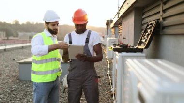 Koruyucu şapkalı çok ırklı inşaat yöneticileri, klima tamiri hakkında modern tabletten bilgi okuyorlar. Sanayi fabrikasının çatısında duran iki adam..