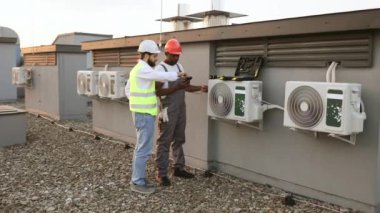 Çatıdaki çok kültürlü inşaat işçileri test araçlarını kullanarak soğutma sistemini birlikte kontrol ediyorlar. Geniş ve gri havalandırma ünitesi birden fazla havalandırma deliği ve borusu var..