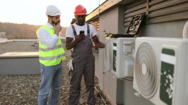 İlgilenen meslektaşım dışarıda modern cihazlara bakarken dikkatli bir işçi dijital çokölçer tutuyor. Çok ırklı tamirciler çatıdaki yeni klimayı tamir ediyor..