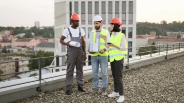 Üç mimardan oluşan hırslı bir ekip çatıdaki çakıl yüzeyinde poz verirken mesafeyi araştırıyor. Çok ırklı meslekdaşlar açık alandaki konut binalarının kopyalarını elinde tutuyor..