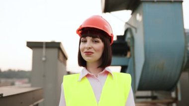 Beyaz kadın portresi, yüksek sarı görünürlüklü yelek ve çatıda duran turuncu koruyucu başlık giyiyor. Mavi gökyüzünün arkaplanı ve büyük klimalı bina.