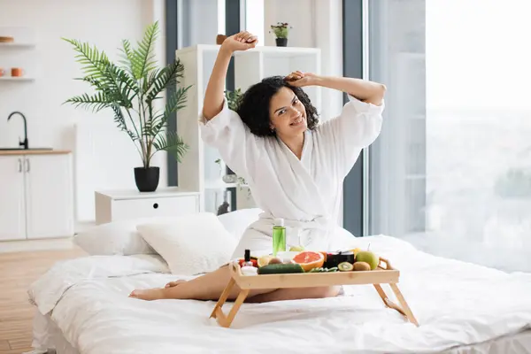 Smiling female in white dressing gown stretching her limbs while prepared ingredients for skin care in bright room of apartment. Joyful housewife increasing life satisfaction by moisturizing her skin.