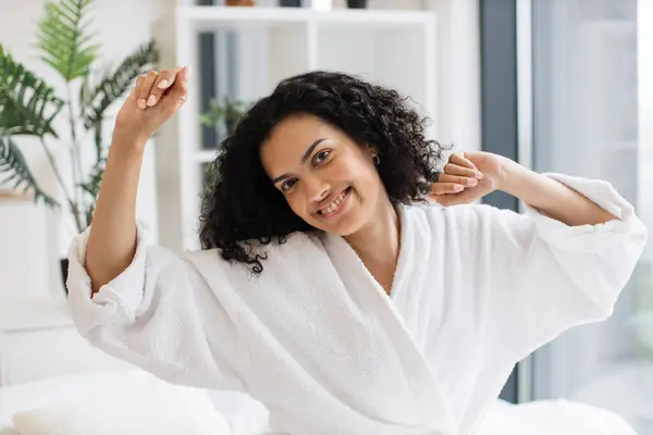 Young female adult in white dressing gown stretching her limbs while resting on cozy bed linen in bright room of studio apartment. Joyful housewife increasing life satisfaction by having fun at home.