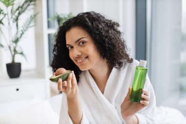 Yatak odasının içinde kozmetik yağ şişesini tutarken elinde avokadoyla kameraya bakan bornozlu güzel sakin bir kadın. Genç bayan yeşil meyvenin büyük cilt faydalarını takdir ediyor..