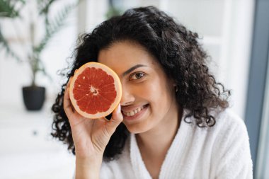 Beyaz bornozlu çekici sakin bir kadın. Bir gözünü greyfurtla kaplıyor. Odanın içi bulanık. Rahatlamış kadın güzellik için C vitamini açısından zengin narenciye uyguluyor..