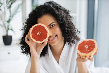 Beyaz bornozlu çekici sakin bir kadın. Bir gözünü greyfurtla kaplıyor. Odanın içi bulanık. Rahatlamış kadın güzellik için C vitamini açısından zengin narenciye uyguluyor..