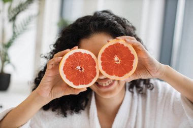 Beyaz bornozlu, çekici sakin bir kadın. Odanın arka planında greyfurt yarısıyla gözlerini kapatıyor. Rahatlamış kadın güzellik için C vitamini açısından zengin narenciye uyguluyor..
