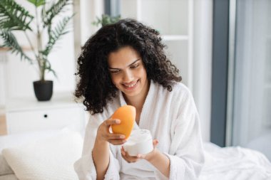 Bornozlu, elinde greyfurt olan ve parlak yatak odasının arka planında sağlıklı cilt için ev yapımı krema yapan mutlu genç bir kadın..