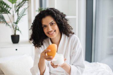 Bornozlu, elinde greyfurt olan ve parlak yatak odasının arka planında sağlıklı cilt için ev yapımı krema yapan mutlu genç bir kadın..