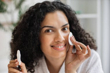 Beyaz bornozlu, pamuk kozmetik süngerler tutan ve arka planda kamera karşısında gülümseyen sağlıklı genç, çok kültürlü bir kadın. Doğal güzellik ve cilt bakımı kavramı.