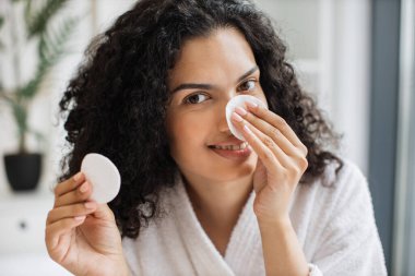 Bornozlu, kıvırcık saçlı, pamuk pedlerle kameraya gülümseyen güzel, pozitif Afrikalı bir kadın burun bölgesinden kozmetik ürünleri alıyor. İnsan kavramı, kozmetik ve cilt bakımı.