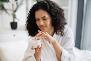 Elinde krem olan ve yüz derisine sürdüğü bornoz kavanozu olan çok kültürlü genç bir kadın. Işıklı yatak odasının arka planında nemlendirici makyaj kremi kullanan bir kadın..