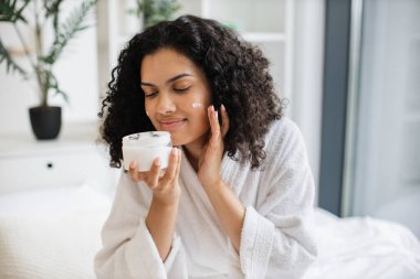 Çekici, çok kültürlü, elinde krema olan ve yüz derisine sürdüğü bornoz kavanozu olan çok kültürlü genç bir kadın. Işıklı yatak odasının arka planında nemlendirici makyaj kremi kullanan bir kadın..