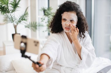 Beyaz bornozlu güzel bir Afrikalı kadın evde kozmetik online video dersinde beyaz bornozlu bir çubukla telefon kullanıyor ve yüzlerine uygulamalı kağıt maske takıyor..