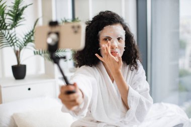 Beyaz bornozlu güzel bir Afrikalı kadın evde kozmetik online video dersinde beyaz bornozlu bir çubukla telefon kullanıyor ve yüzlerine uygulamalı kağıt maske takıyor..