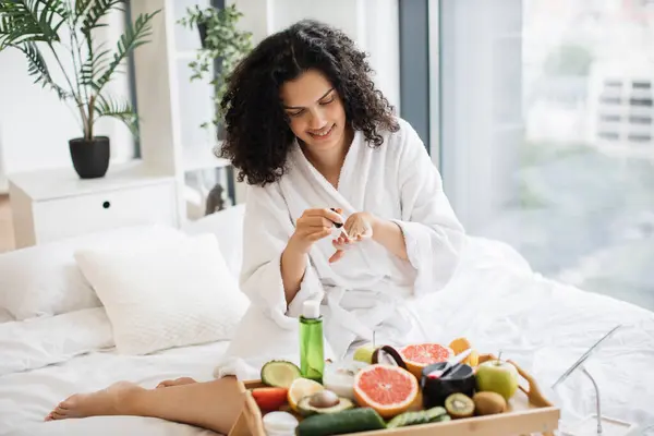 Smiling female in white bathrobe making manicure while prepared ingredients for skin care in bright bedroom of apartment. Joyful housewife increasing life satisfaction by paints nails with polish.