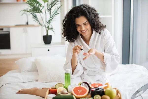 Portrait of female in white bathrobe making manicure while prepared ingredients for skin care in bright bedroom of apartment. Joyful housewife increasing life satisfaction by paints nails with polish.