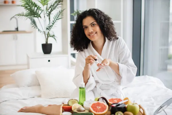 Multiracial female in white bathrobe making manicure while prepared ingredients for skin care in bright bedroom of apartment. Joyful housewife increasing life satisfaction by paints nails with polish.