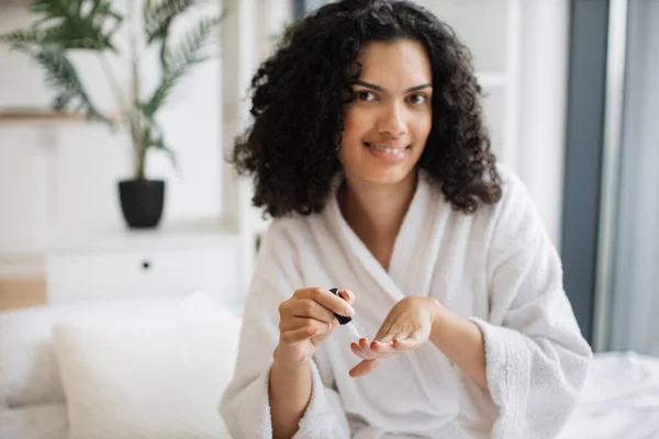 Multiracial female in white bathrobe making manicure while prepared ingredients for skin care in bright bedroom of apartment. Joyful housewife increasing life satisfaction by paints nails with polish.