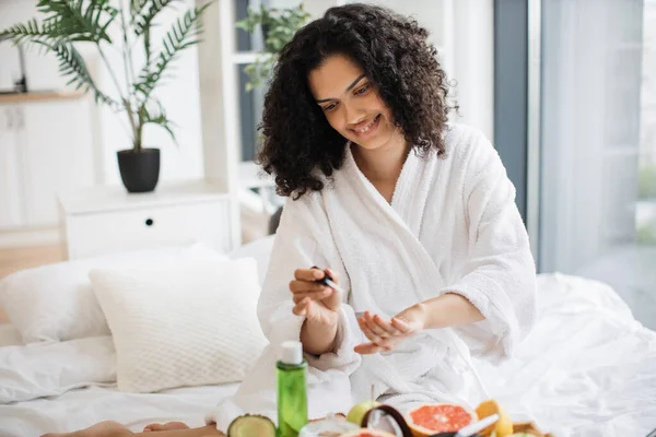 Portrait of female in white bathrobe making manicure while prepared ingredients for skin care in bright bedroom of apartment. Joyful housewife increasing life satisfaction by paints nails with polish.