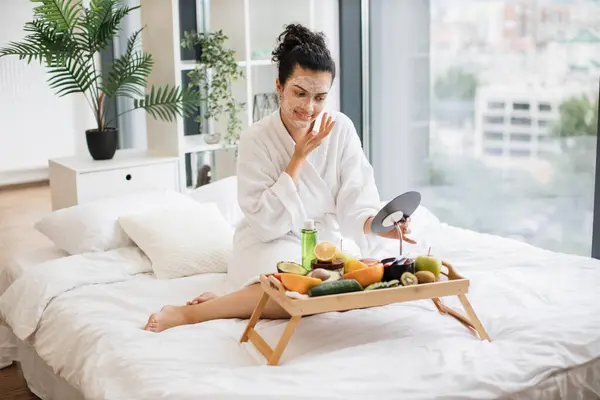 Relaxed adult person in after-shower wear applying facial mask using makeup mirror from tray table while sitting on bed in studio room. Serene lady moisturising skin with fruits and cosmetics.